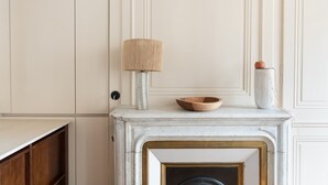 Ein Zimmer mit Kamin, einer Holzschale auf dem Kaminvorsprung, einer Lampe und einer Vase.