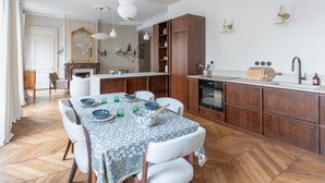 Ein Essbereich mit einem Tisch für vier Personen, Holzschränke, eine Küche mit Spüle und Herd sowie ein großer Spiegel über dem Kamin.