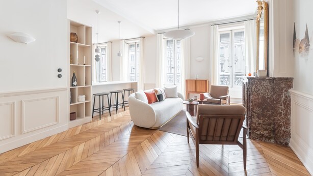 Ein großzügiger Wohnraum mit Fischgrätparkett, einer Couch, einem Sessel, einem Kamin und einem Bücherregal.