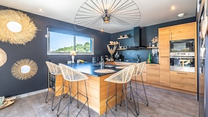 A modern kitchen with a central island, bar stools, wooden cabinets, and a large window. - Villa Mindoya - Villa pour 10, piscine chauffée (Labenne)