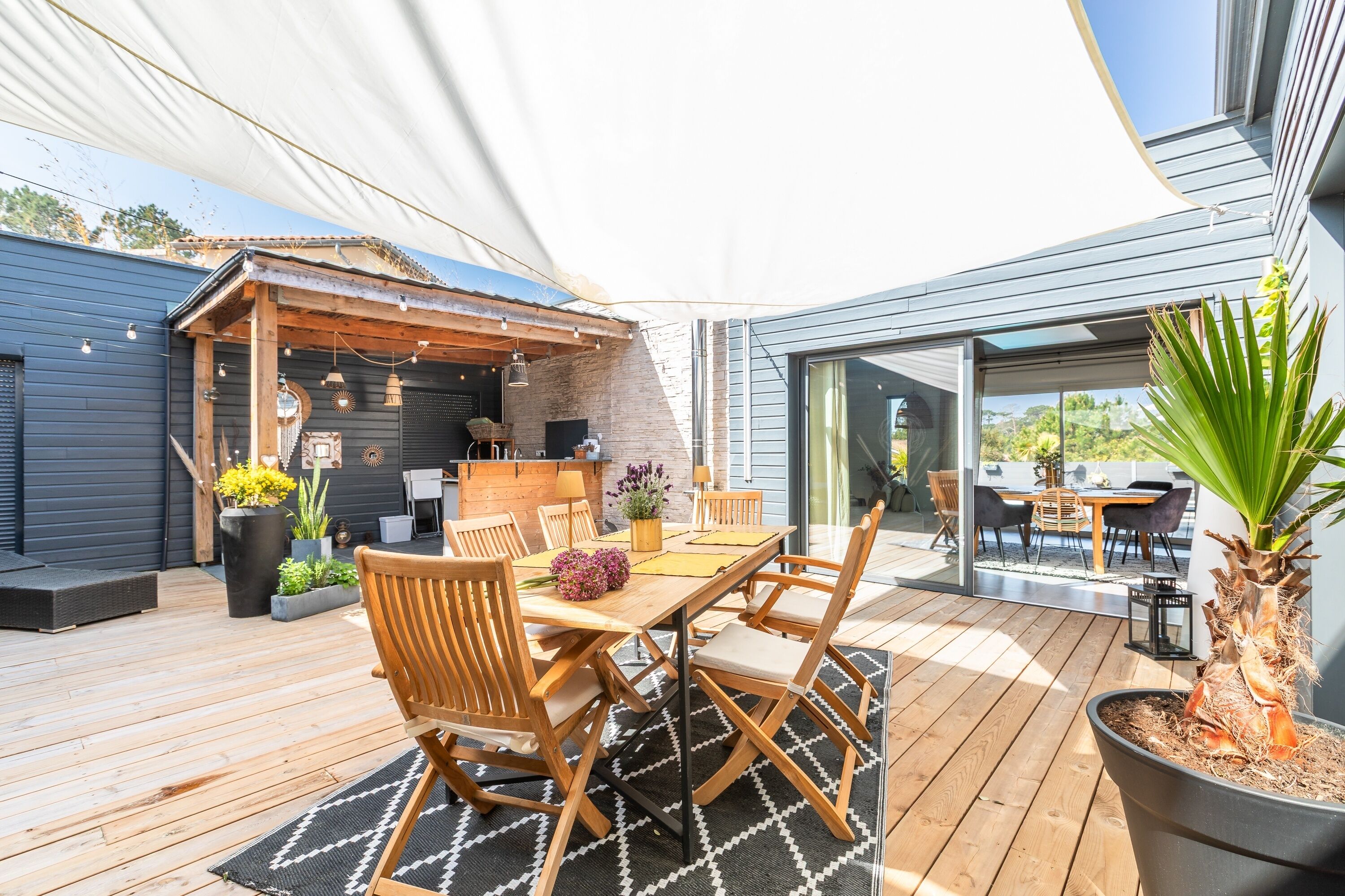 Een terras buiten met een houten tafel en stoelen, omringd door potplanten en een pergola.