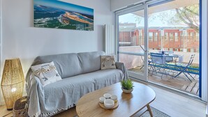 Ein modernes Wohnzimmer mit einem grauen Sofa, einem Couchtisch mit Tassen, einem großen Fenster mit Blick auf einen Balkon und einem Landschaftsbild.