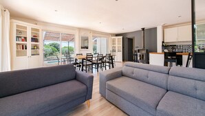 Un salon moderne avec des canapés gris, un coin repas avec une table et des chaises en bois, et une cuisine avec des meubles blancs et un plan de travail noir.