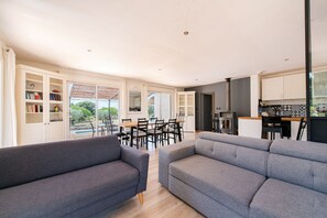 Ein modernes Wohnzimmer mit grauen Sofas, ein Essbereich mit einem Holztisch und Stühlen sowie eine Küche mit weißen Schränken und einer schwarzen Spritzschutzwand.