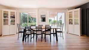Une salle à manger avec une grande table et des chaises, entourée de meubles blancs aux portes vitrées, de grandes fenêtres et offrant une vue sur la verdure à l’extérieur.