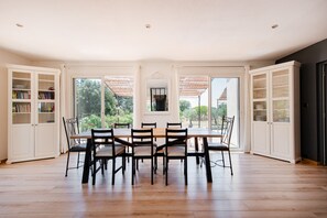 Ein Esszimmer mit großem Tisch und Stühlen, umgeben von weißen Schränken mit Glastüren, großen Fenstern und Blick auf Grünflächen.