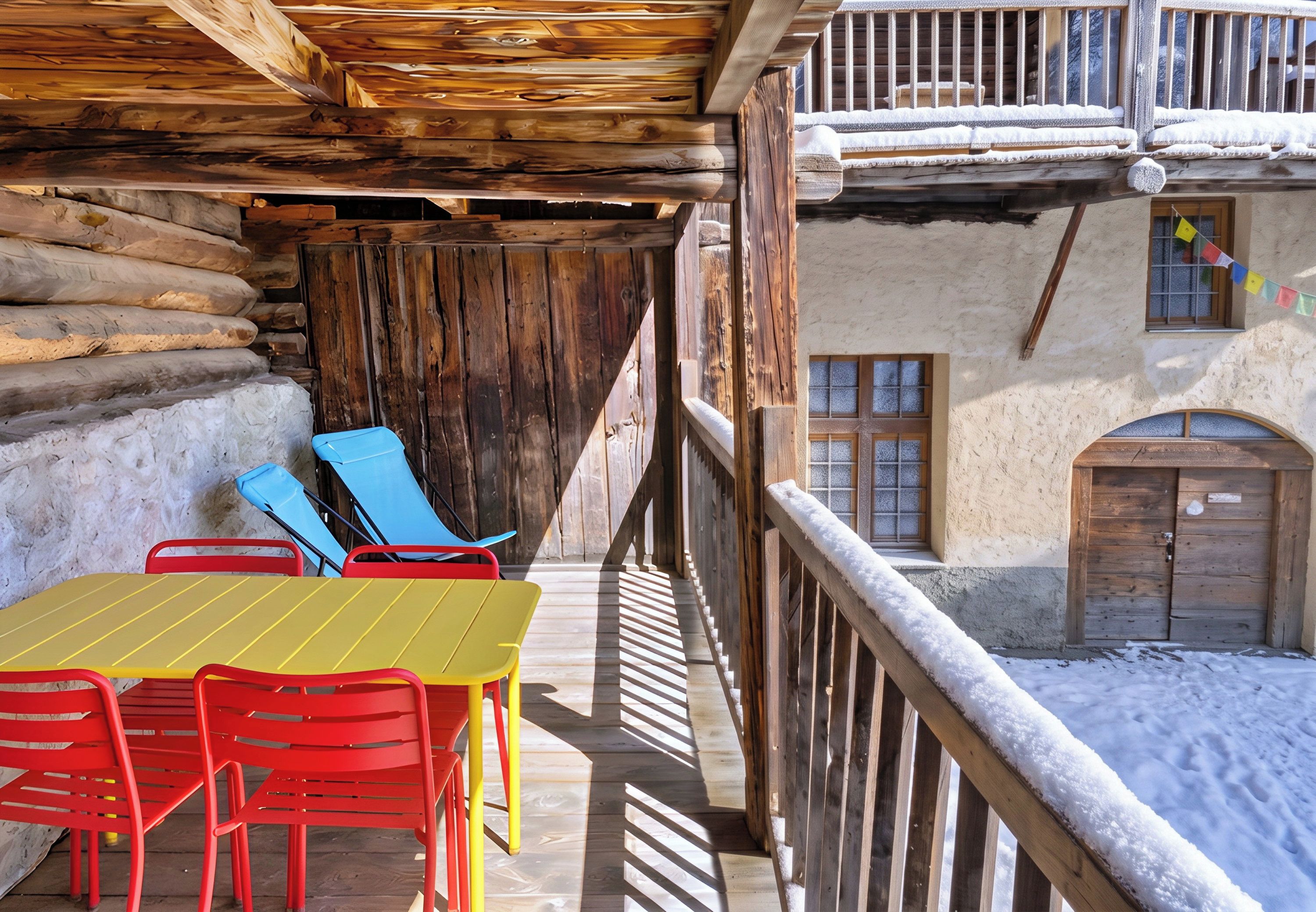 A wooden balcony with a table and chairs, a snow-covered ground, and a building with a window and a door.