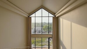 Ein Zimmer mit großem Fenster, schrägem Dach und Blick auf eine hügelige Landschaft.