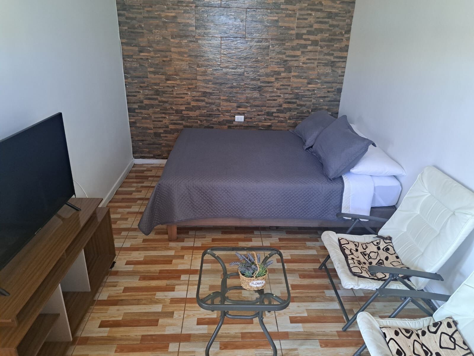 A small, neatly arranged bedroom with a bed, a TV, a chair, and a small table.
