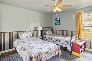 A bedroom with two beds, a ceiling fan, a window with curtains, and a small table with a lamp.