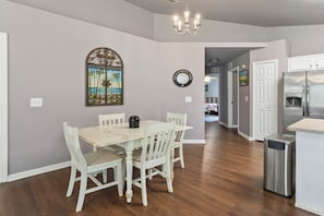 A dining area with a table and chairs, a chandelier, and a kitchen with a refrigerator and trash can.