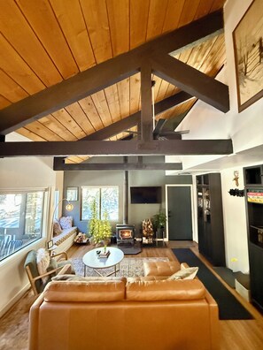 A spacious living room with a high ceiling, a fireplace, a sofa, a coffee table, and a television. - Dreamy: Modern + Unique + Garage + HotTub - Walk to Village (Mammoth Lakes)
