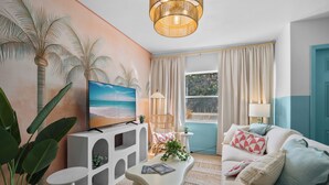 A living room with a TV, a white sofa, a coffee table, and a painting of palm trees. - Stylish 5min Downtown Orlando (Orlando)