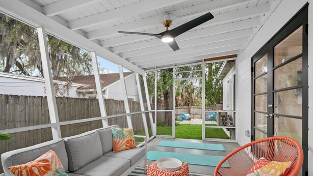 A covered patio with a white ceiling, a gray sofa with patterned cushions, a red chair, and a view of a backyard. - Stylish 5min Downtown Orlando (Orlando)