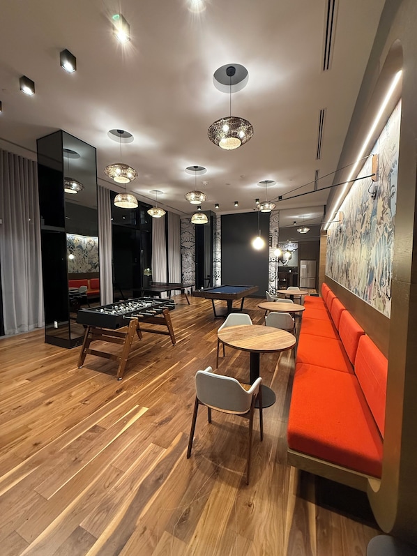 A modern lounge area with wooden flooring, a foosball table, multiple seating options, and decorative lighting fixtures.
