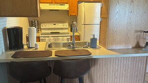 Una cucina compatta con mobili in legno, un frigorifero bianco, un piano cottura e due sgabelli da bar.