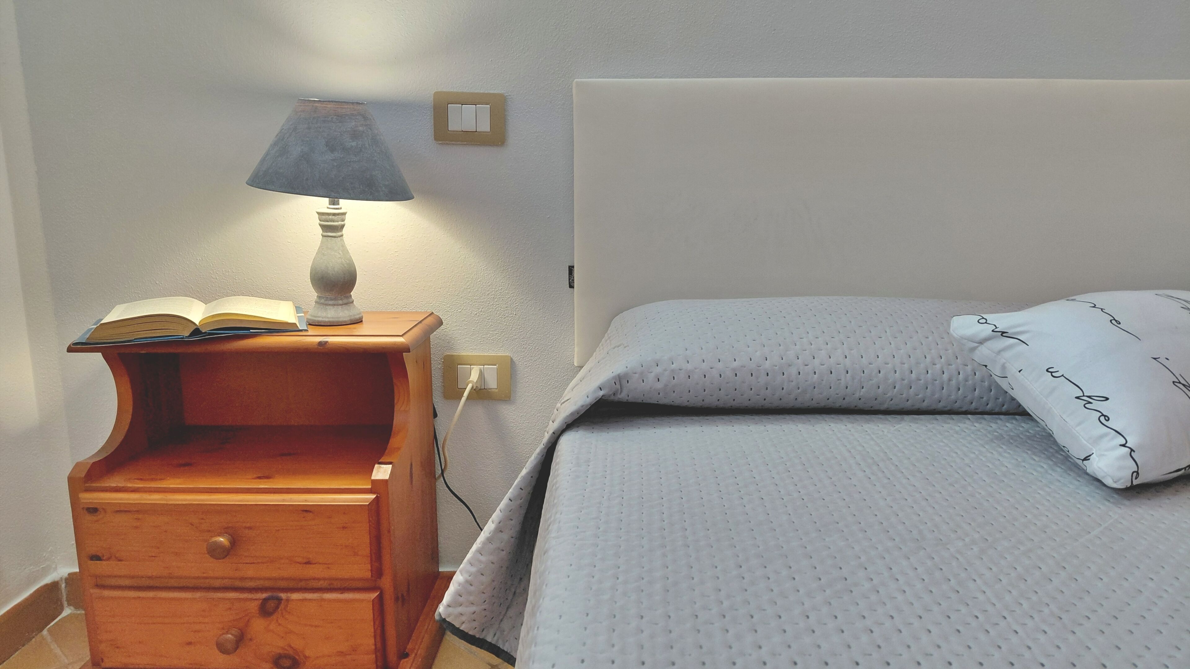 A bed with a pillow and a bedside table with a lamp and an open book.