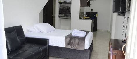 A compact room with a bed, a sofa, a small kitchenette, and a TV mounted on the wall.