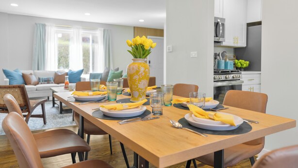 Un coin repas avec une table en bois dressée pour quatre personnes, des chaises, un vase rempli de fleurs jaunes et, au fond, une cuisine.
