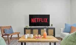 A living room with a television displaying 'NETFLIX,' a wooden coffee table with a plate of food and drinks, a wicker chair, and a sofa.
