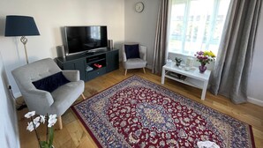 A living room with a flat-screen TV, a grey sofa, a white side table, a red patterned rug, and a window with curtains. - Entire Home. 3 bedroom house in Edinburgh suburb (Edinburgh)