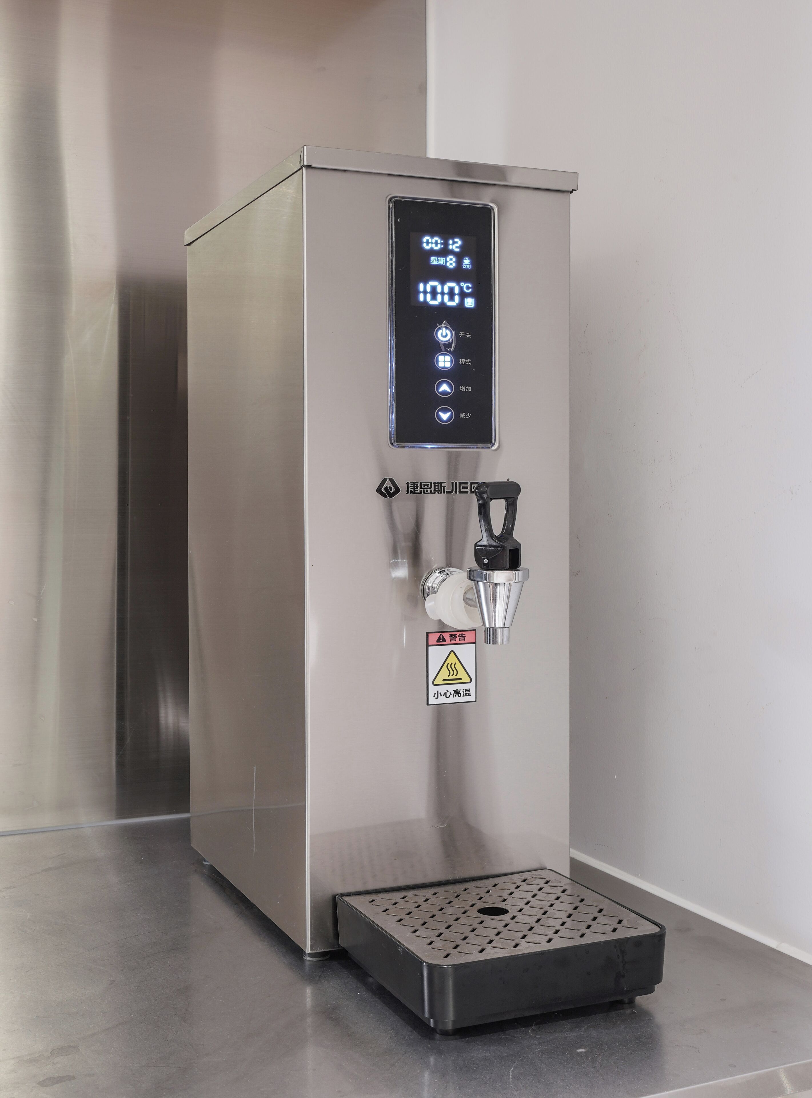 A stainless steel water dispenser with a digital display, a warning label, and a drainage grate.