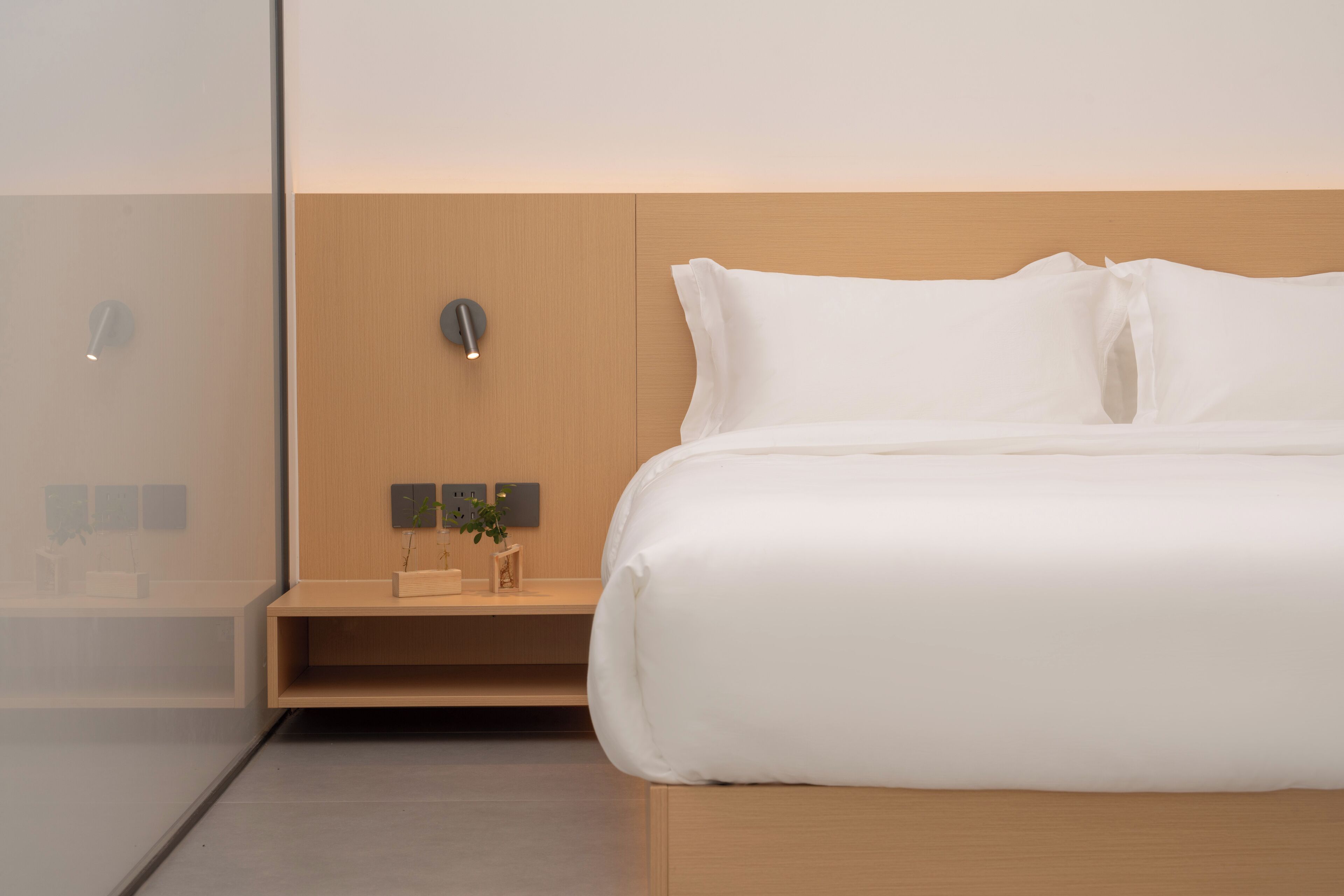 Una habitación de hotel moderna con cabecera de madera, una mesita de noche con una planta y una cama grande con ropa blanca.
