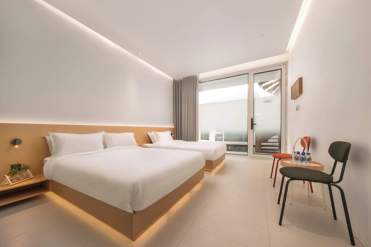 A modern hotel room with two beds, a desk, a chair, and a view of the outside.