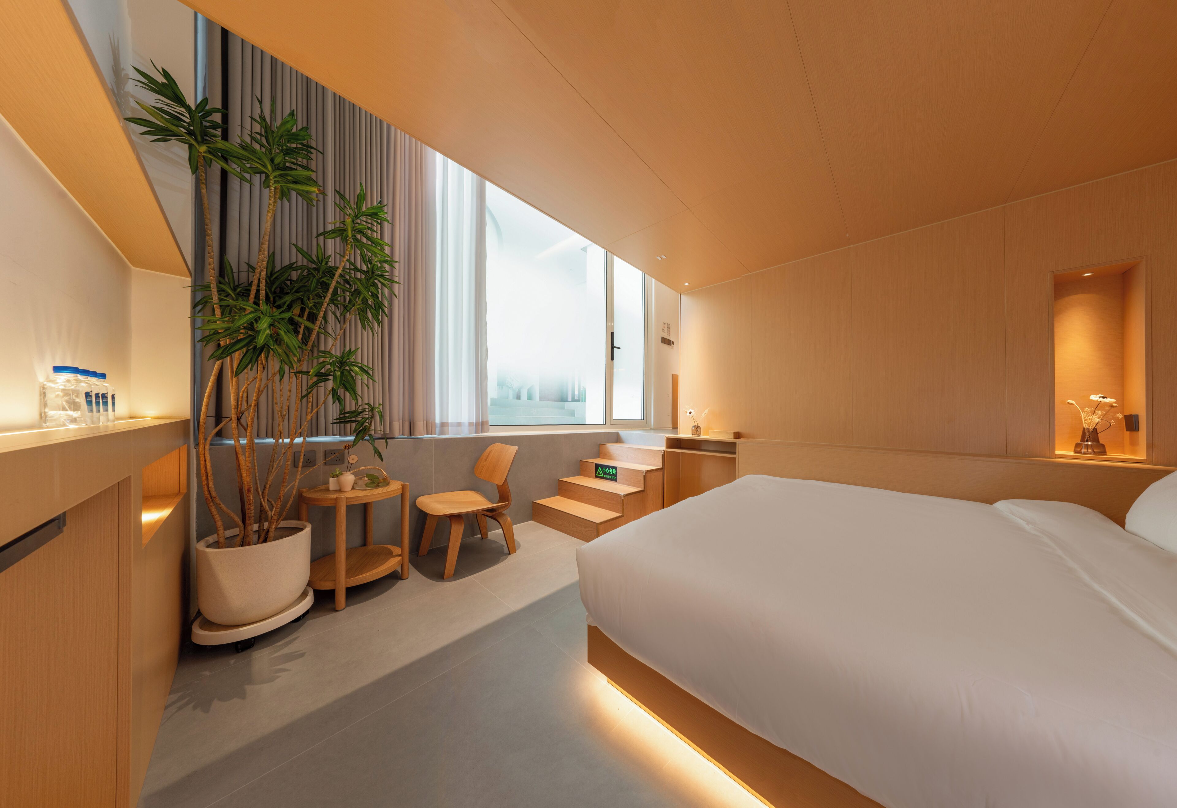 A modern hotel room with a large bed, a seating area with a chair and a small table, a potted plant, and a view of the city through large windows.