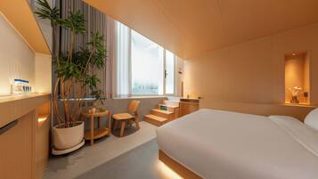 A modern hotel room with a large bed, a seating area with a chair and a small table, a potted plant, and a view of the city through large windows.