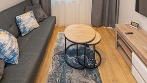 A modern living room with a grey sofa, a round wooden table, a floor lamp, and a large window with grey curtains.