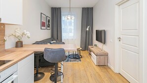 A compact living space with a kitchenette, a dining area, a seating area with a sofa, a TV mounted on the wall, and a large window with curtains.