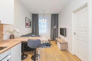 A compact living space with a kitchenette, a dining area, a seating area with a sofa, a TV mounted on the wall, and a large window with curtains. - Prime City 1BD Apartment by Domvia (Kaunas)