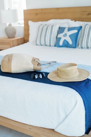 Ein ordentlich bezogenes Bett mit einem blau-weiß gestreiften Kopfkissen, einem beigen Kopfkissen und einem Strohhut auf einer blauen Decke.