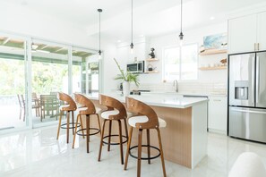 A modern kitchen with a central island, bar stools, a stainless steel refrigerator, and a large window providing natural light. - NEW Luxury Grace Bay villa,Pool,2 min>Beach&Reef (The Bight Settlement)