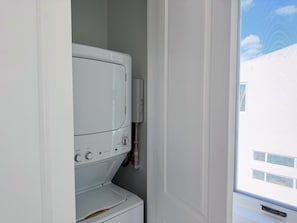 A compact laundry room with a front-loading washer and dryer, a water heater, and a window with a view of buildings outside. - NEW Luxury Grace Bay villa,Pool,2 min>Beach&Reef (The Bight Settlement)