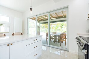 A modern kitchen with white cabinetry, a marble countertop, and a view of an outdoor patio with wooden furniture. - NEW Luxury Grace Bay villa,Pool,2 min>Beach&Reef (The Bight Settlement)