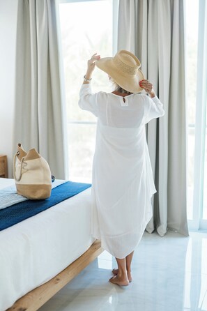 A person in a white robe standing in a room with a bed, a wooden floor, and a window with curtains. - NEW Luxury Grace Bay villa,Pool,2 min>Beach&Reef (The Bight Settlement)