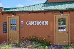 A building labeled 'GAME ROOM' with two doors, a signboard with operating hours, and a small garden area with plants and flowers. - The Covered Corral Luxury RV Lot River Ranch 253 (Lake Wales)