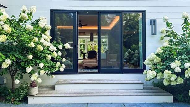 Een modern huis met grote glazen deuren, een houten terras en struiken witte hortensia's.