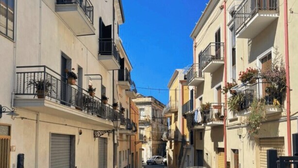 Una estrecha calle adoquinada flanqueada por edificios de varios pisos, cada uno con balcones y macetas.