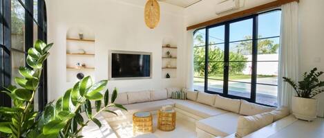 A spacious living room with a large sectional sofa, a flat-screen TV, and a large window offering a view of greenery.