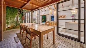 An outdoor wooden deck with a dining table and chairs, large glass doors leading to an interior space, and a ceiling with wooden slats. - Villa Yasmine Althea by Senada (Pecatu)