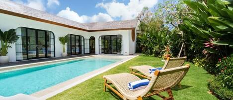 A modern house with a swimming pool, two wicker lounge chairs, and a well-maintained garden.