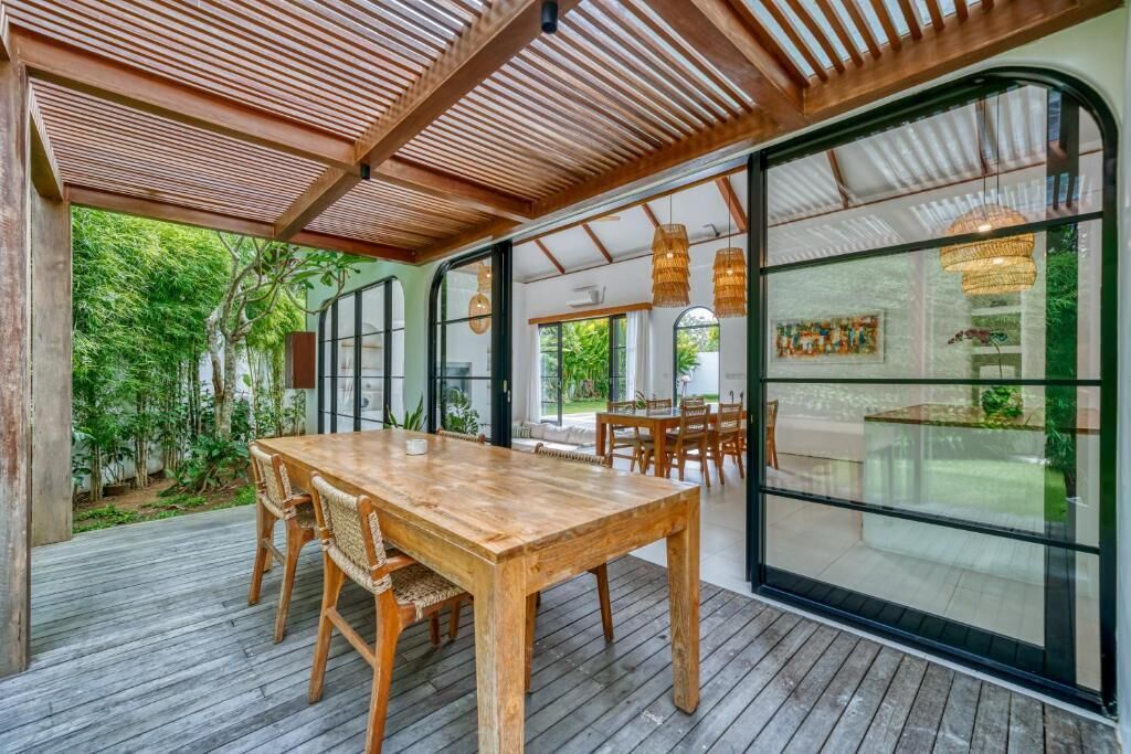 Een houten terras met een eettafel en stoelen, grote glazen deuren en uitzicht op groen.