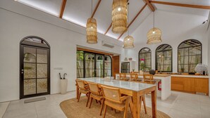 A spacious dining area with a large table and chairs, a kitchen area with wooden cabinets, and large windows with a view of greenery. - Villa Yasmine Althea by Senada (Pecatu)