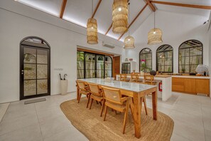 A spacious dining area with a large table and chairs, a kitchen area with wooden cabinets, and large windows with a view of greenery. - Villa Yasmine Althea by Senada (Pecatu)