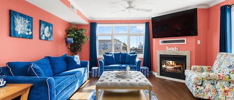 Una sala de estar con chimenea, dos sofás azules, un sillón colorido y un ventanal grande.