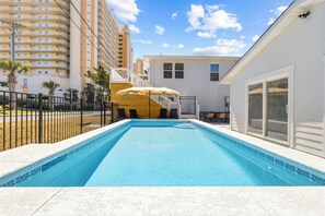 Piscina com uma casa branca ao fundo, uma área cercada e um edifício alto à esquerda.