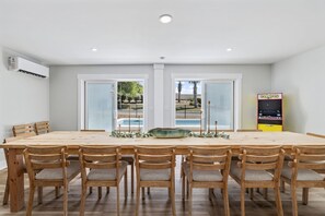 A dining area with a large wooden table and chairs, a decorative bowl, and a Pac-Man arcade machine. - Sea Breeze - 6BR Coastal Escape w/ Heated Pool + Dual Kitchens (Walk to Beach!) (North Myrtle Beach)